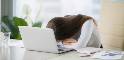 Mulher jovem cansada e com a cabeça apoiada na mesa em frente ao notebook