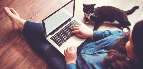 Mulher jovem sentada no chão com notebook no colo e gato ao lado