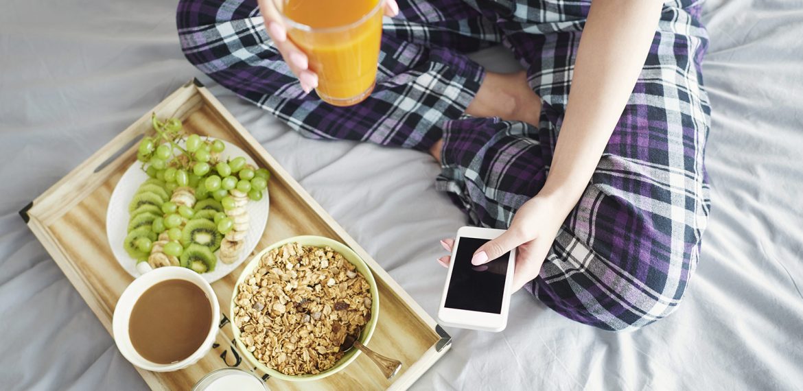 Mulher toma café da manhã com frutas, cereal, café e suco ao mesmo tempo que usa celular